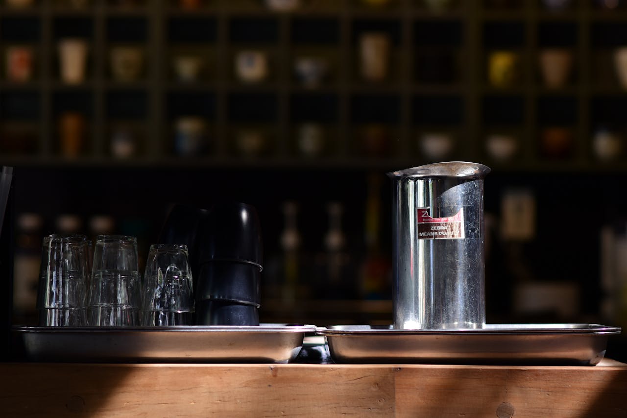 Elegant arrangement of glassware and a metal pitcher on a wooden counter in a dimly lit cafe setting.